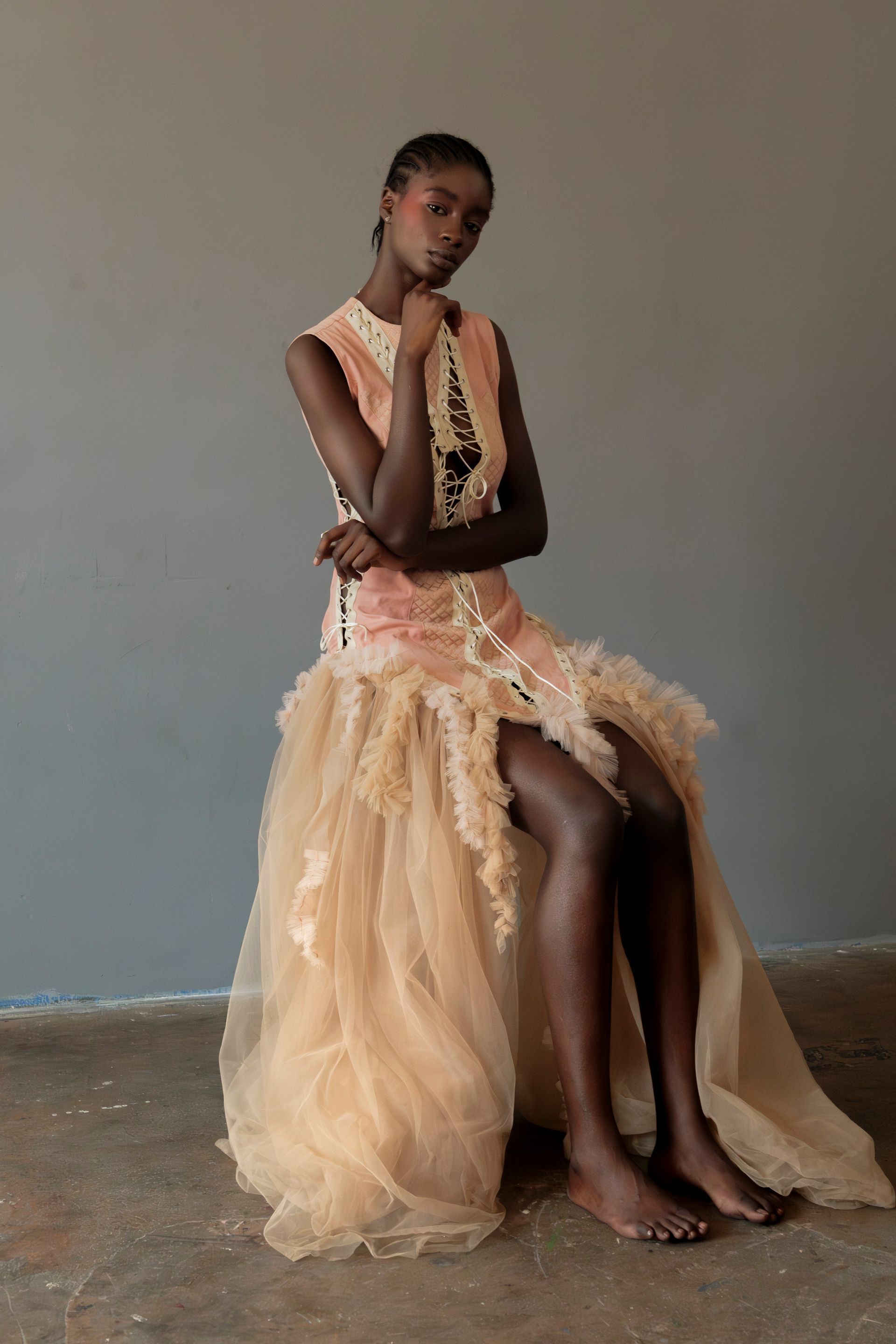 Una donna con un abito color pesca è seduta, con la mano sul mento, davanti a un muro grigio.