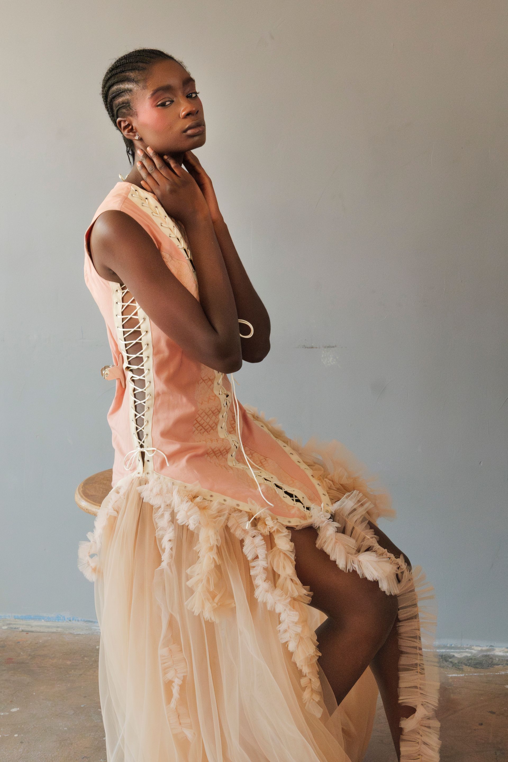 Donna con abito color pesca e gonna in tulle, seduta con le mani sul collo. Capelli intrecciati e trucco rosa.