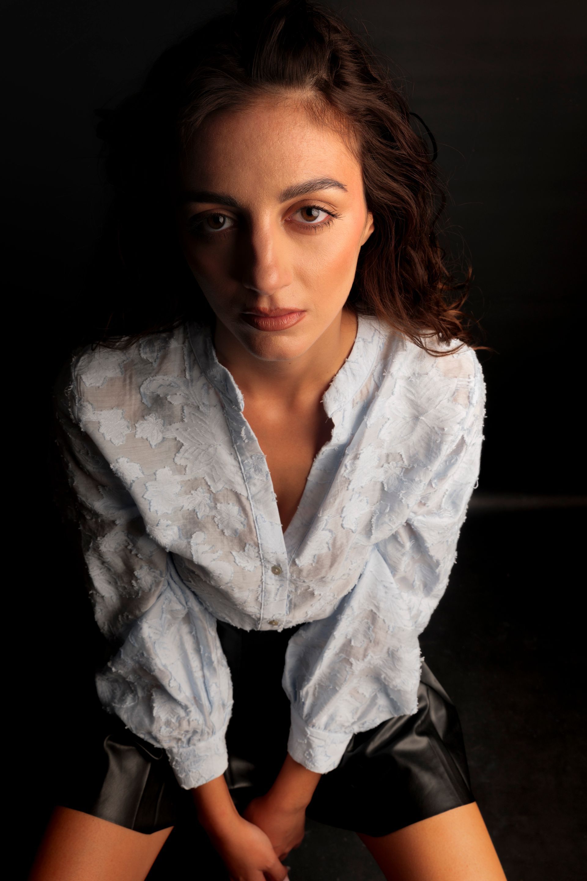 Donna con i capelli ricci, con una camicia azzurra ricamata e pantaloncini neri, che guarda la telecamera.