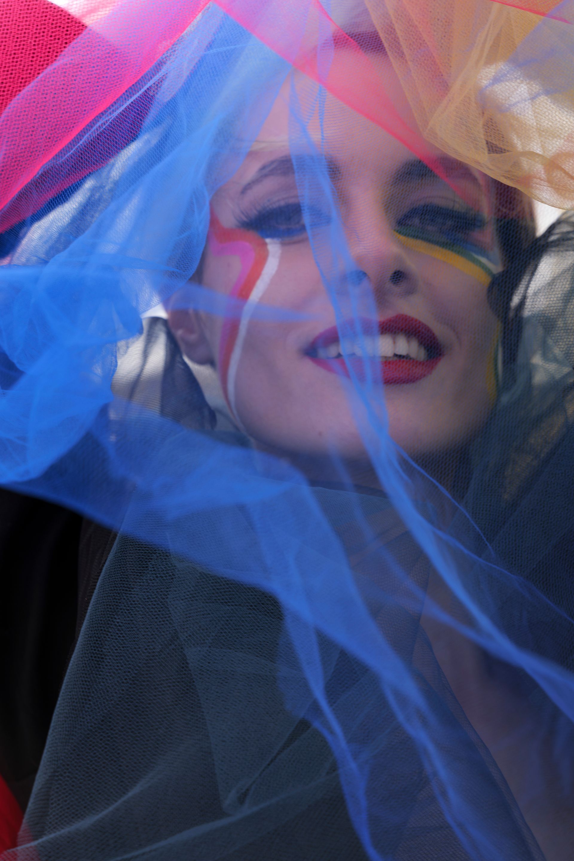 Una donna con il viso dipinto di colori vivaci sorride attraverso il tulle blu.
