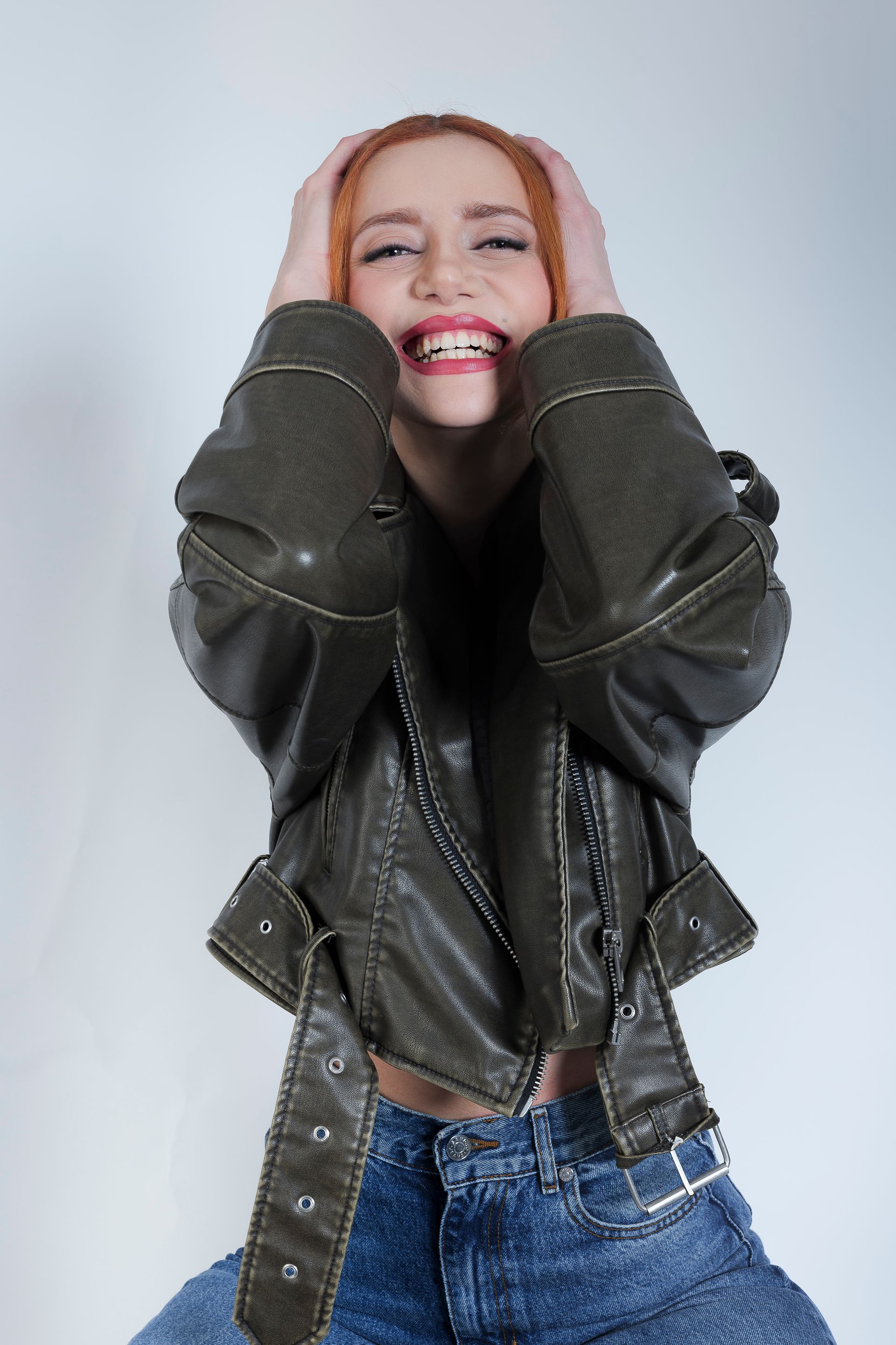 Donna con i capelli rossi, giacca di pelle verde e jeans blu, che ride con le mani sulla testa.