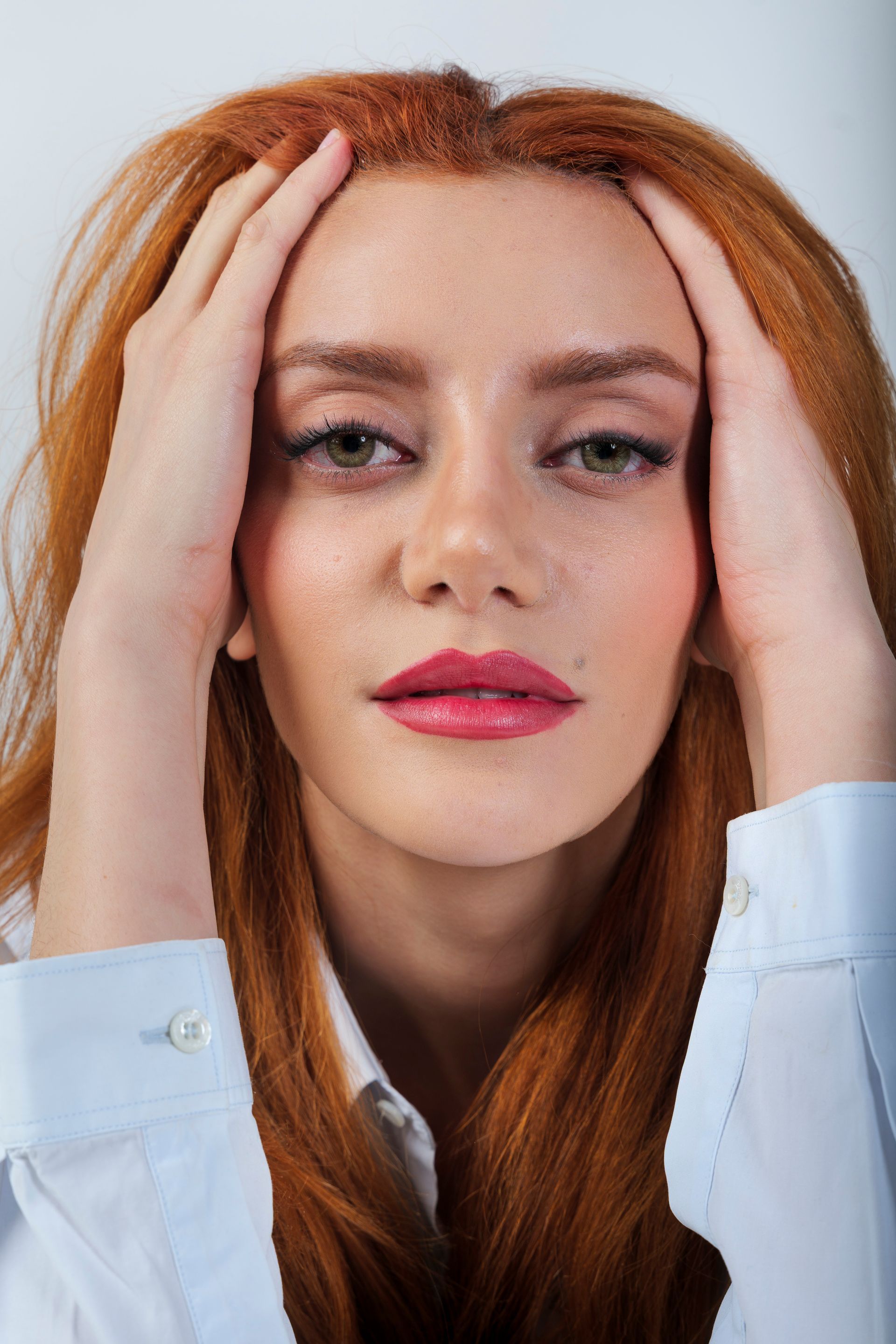 Donna con i capelli rossi, mani sulla testa, che indossa una camicia bianca.
