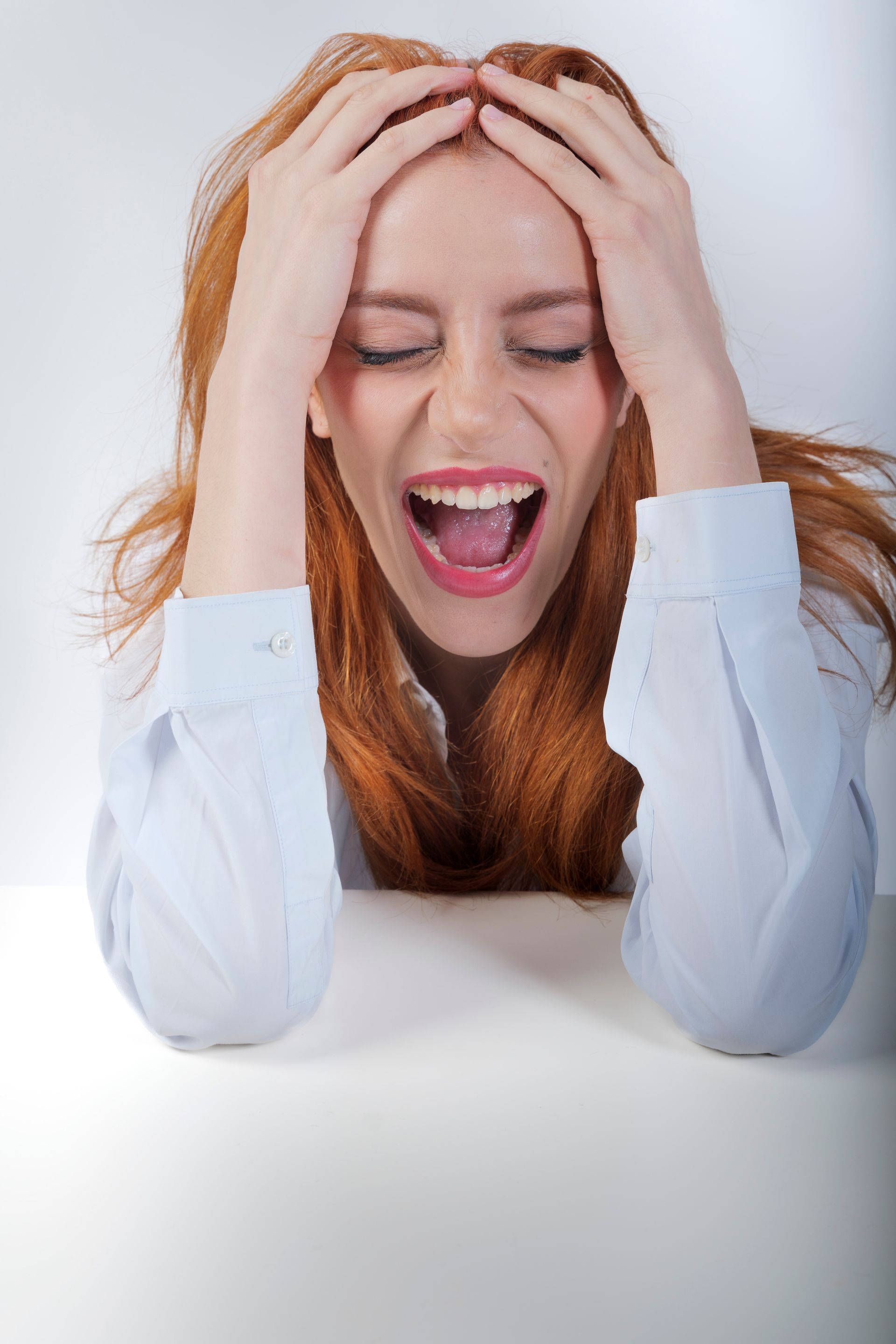 Donna con i capelli rossi, occhi chiusi, mani sulla testa, che urla per la frustrazione.