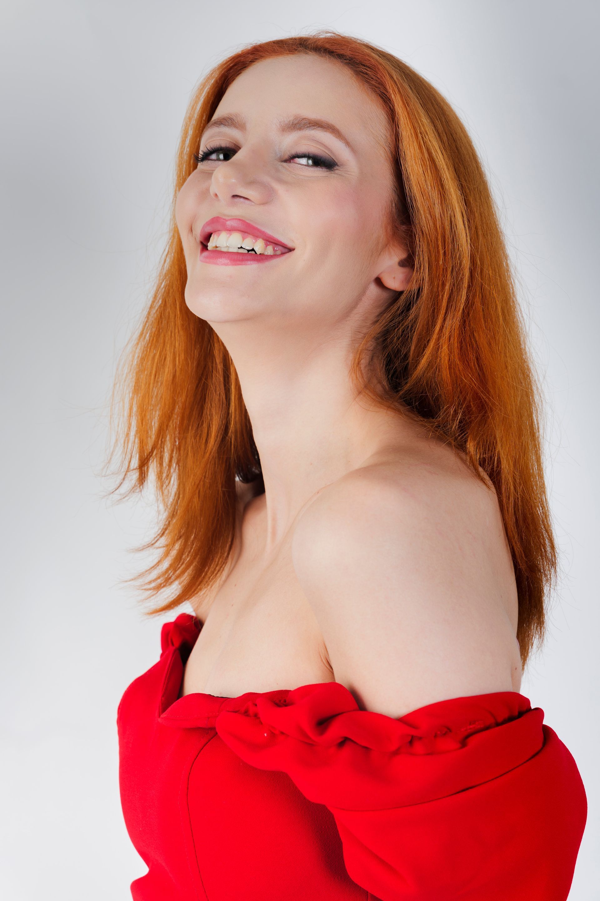 Una donna con i capelli rossi sorride, indossando un abito rosso con le spalle scoperte, su sfondo bianco.