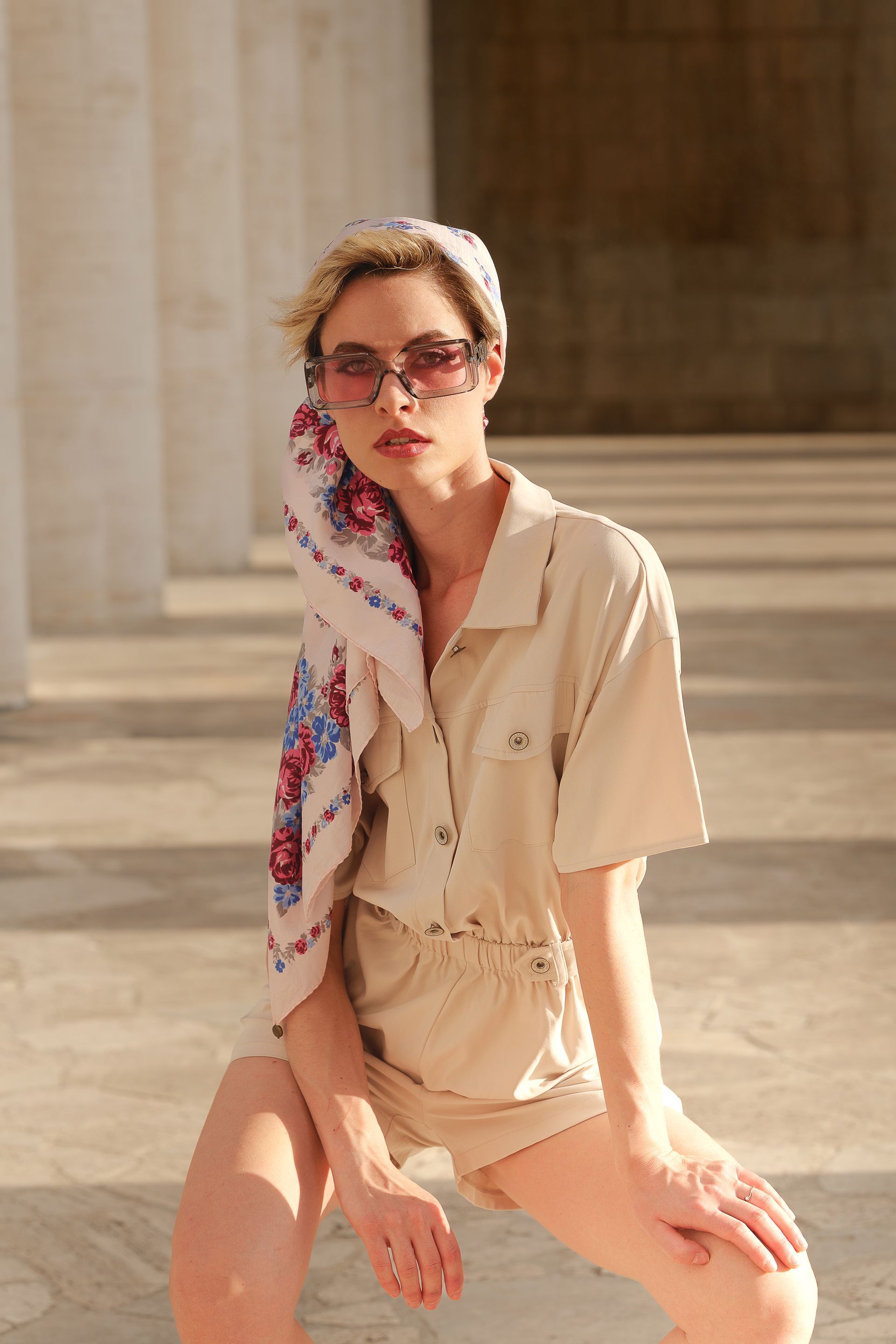 Donna con foulard e occhiali da sole, in una tuta color sabbia, seduta all'aperto vicino alle colonne.