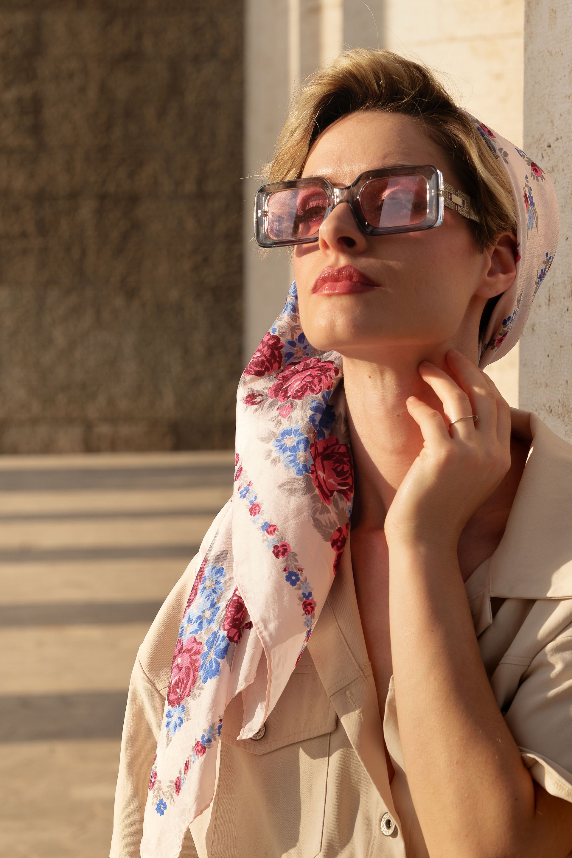 Donna con occhiali da sole e foulard, illuminata dalla luce del sole, appoggiata a un edificio.