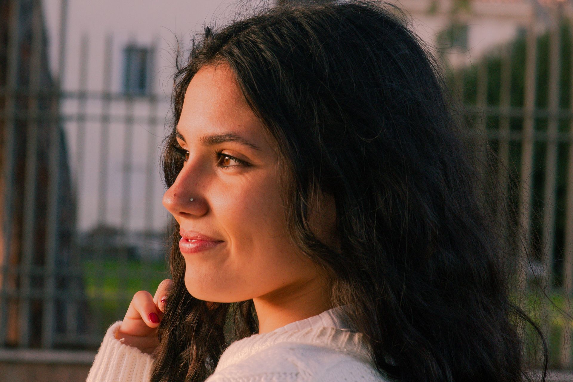 Donna con capelli scuri e ondulati, sorridente, che guarda di lato in un ambiente tranquillo e all'aperto, con una recinzione.