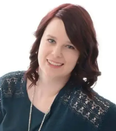 Woman with auburn hair smiles, wearing a teal top with lace sleeves, and a silver necklace, against a white background.