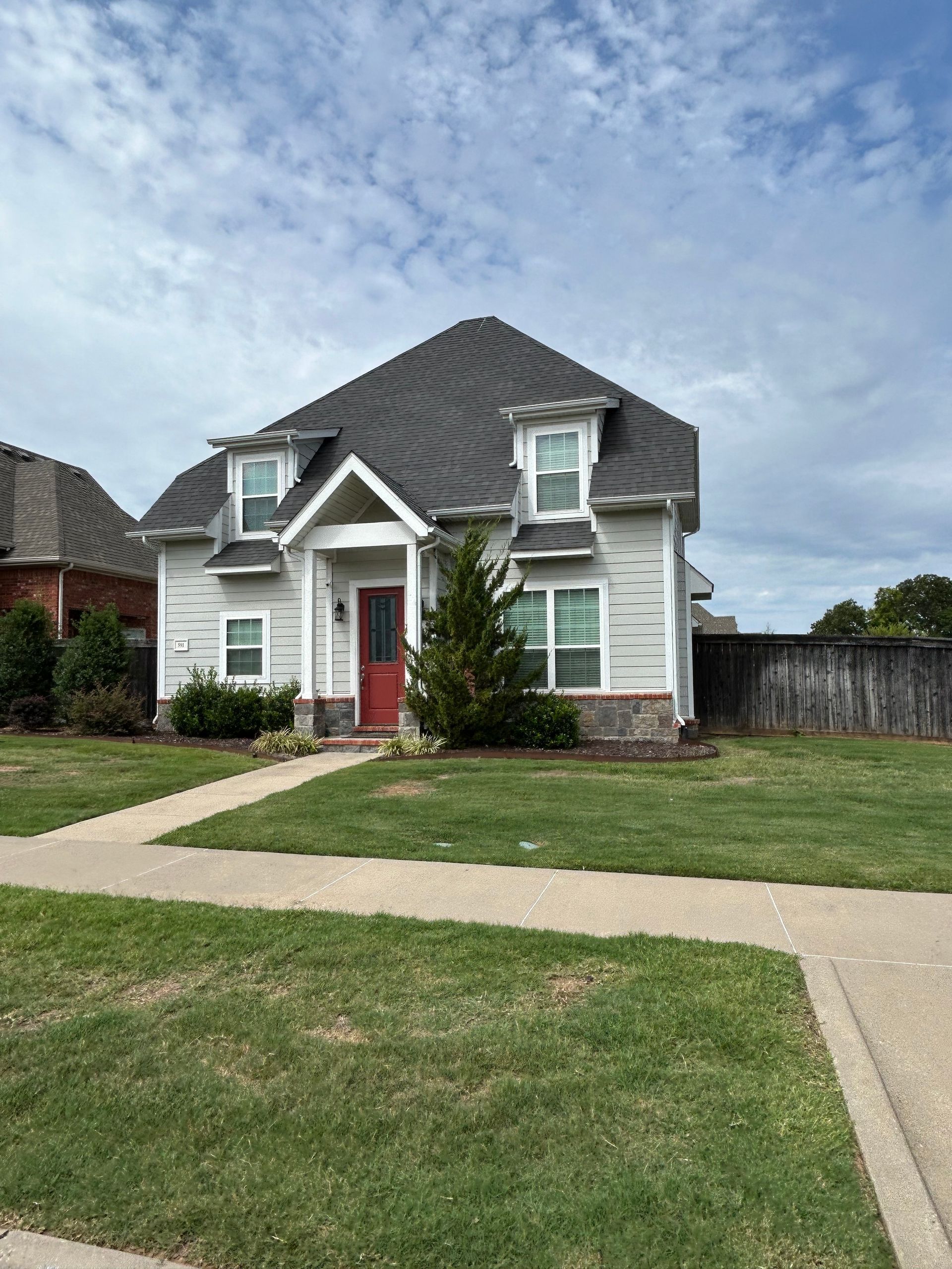 Two-story house with gray siding, red front door, dormers, and dark roof on a green lawn.