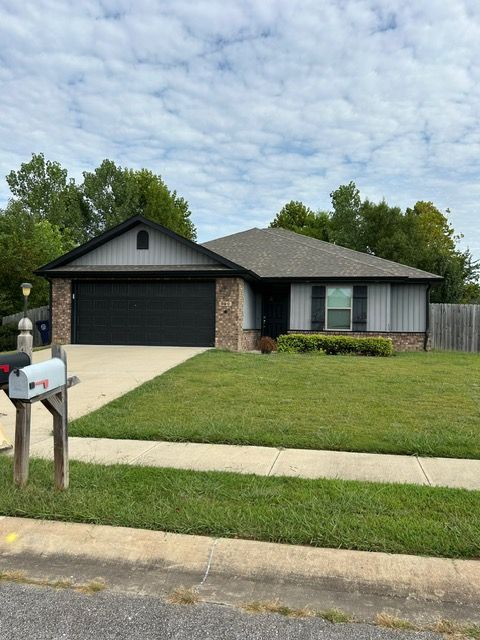 Ranch-style house with gray siding, brick accents, black garage door, and green lawn. A mailbox is in the foreground.
