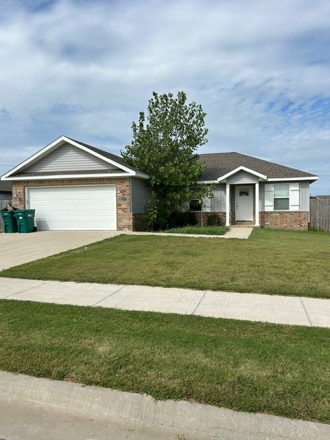 Single-story house with gray siding, brick accents, and a white garage door. Green lawn and a small tree. Overcast sky.