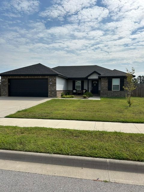 Single-story house with a black garage door and brick and white siding, sitting on a grassy lawn with a blue sky.