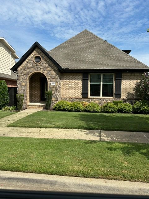 Tan brick and stone house with dark shutters and brown roof, set on a green lawn.