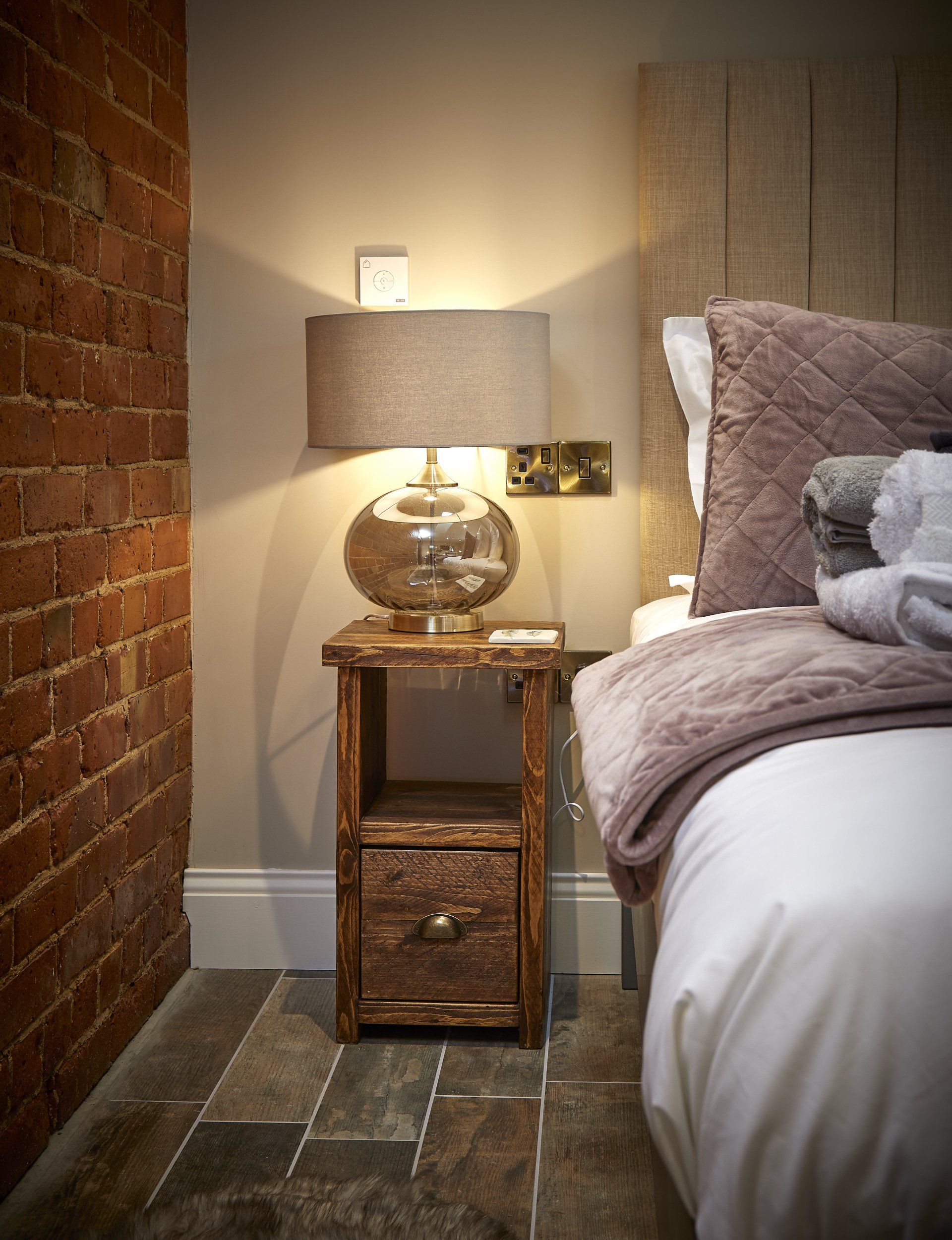A bedroom with a bed , nightstand , lamp and brick wall.