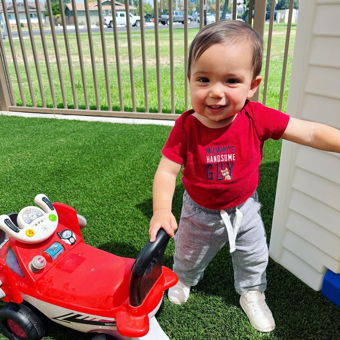 A baby is standing next to a red toy car.