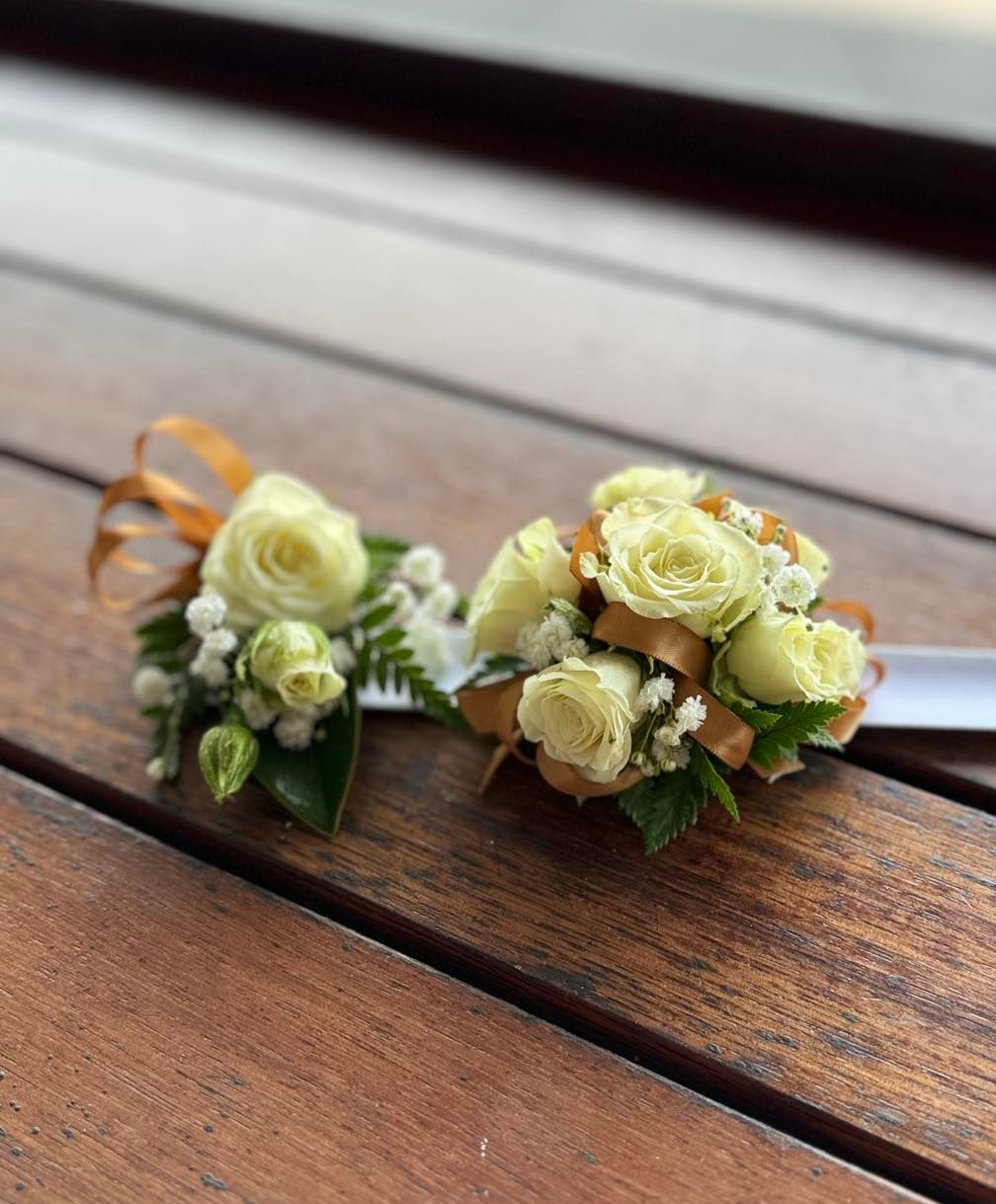 Two wrist corsages with cream-colored roses, baby's breath, and greenery on a wooden surface - J'adore Flowers & Gifts Cairns in Earlville, QLD