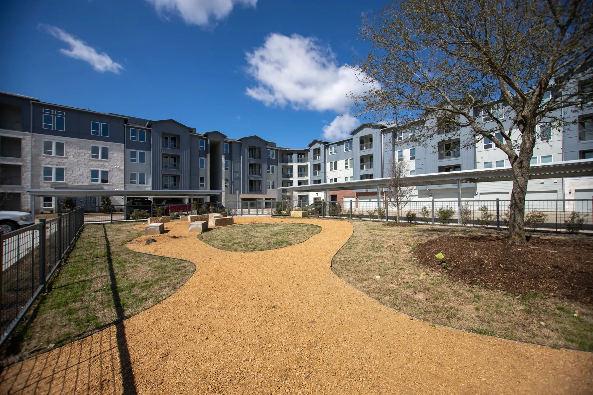 Exterior view of an apartment community courtyard with a curved gravel path, benches, and surrounding buildings.