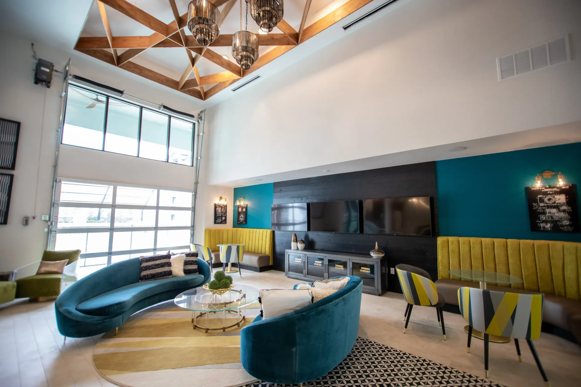 Spacious modern resident lounge with teal curved sofas, yellow banquettes, glass coffee tables, and wall TVs.