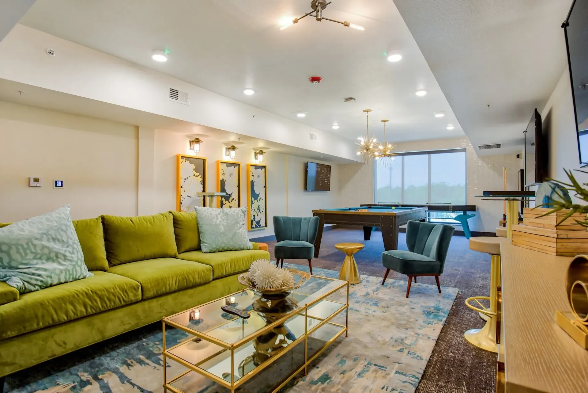 Indoor community lounge with a green sofa, teal chairs, glass coffee table, and a pool table.