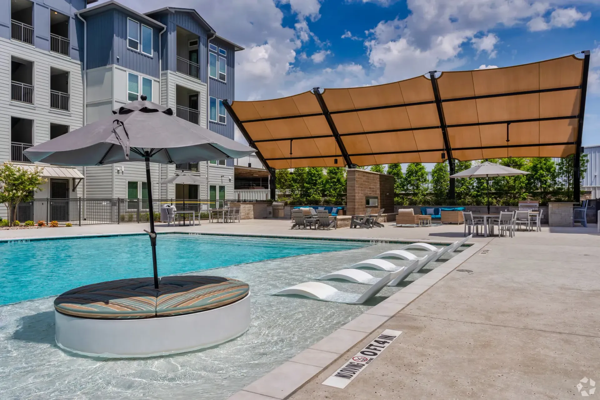 Outdoor apartment pool area with umbrellas, loungers, and a large shade canopy.