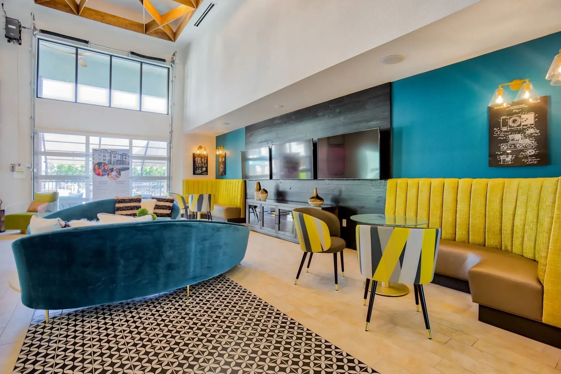 Bright communal lounge with teal accent wall, yellow banquettes, a blue curved sofa, and wall-mounted TVs.
