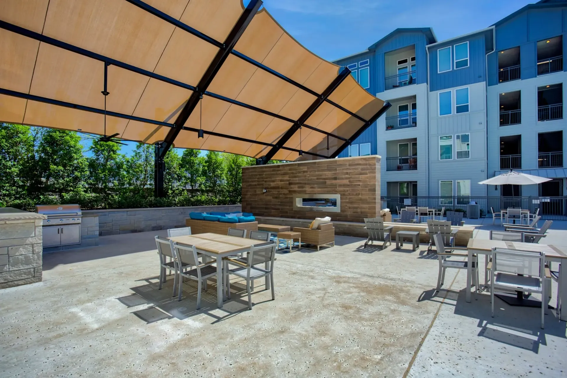 Outdoor communal patio with seating, a shade canopy, grill, and modern apartment building in the background.