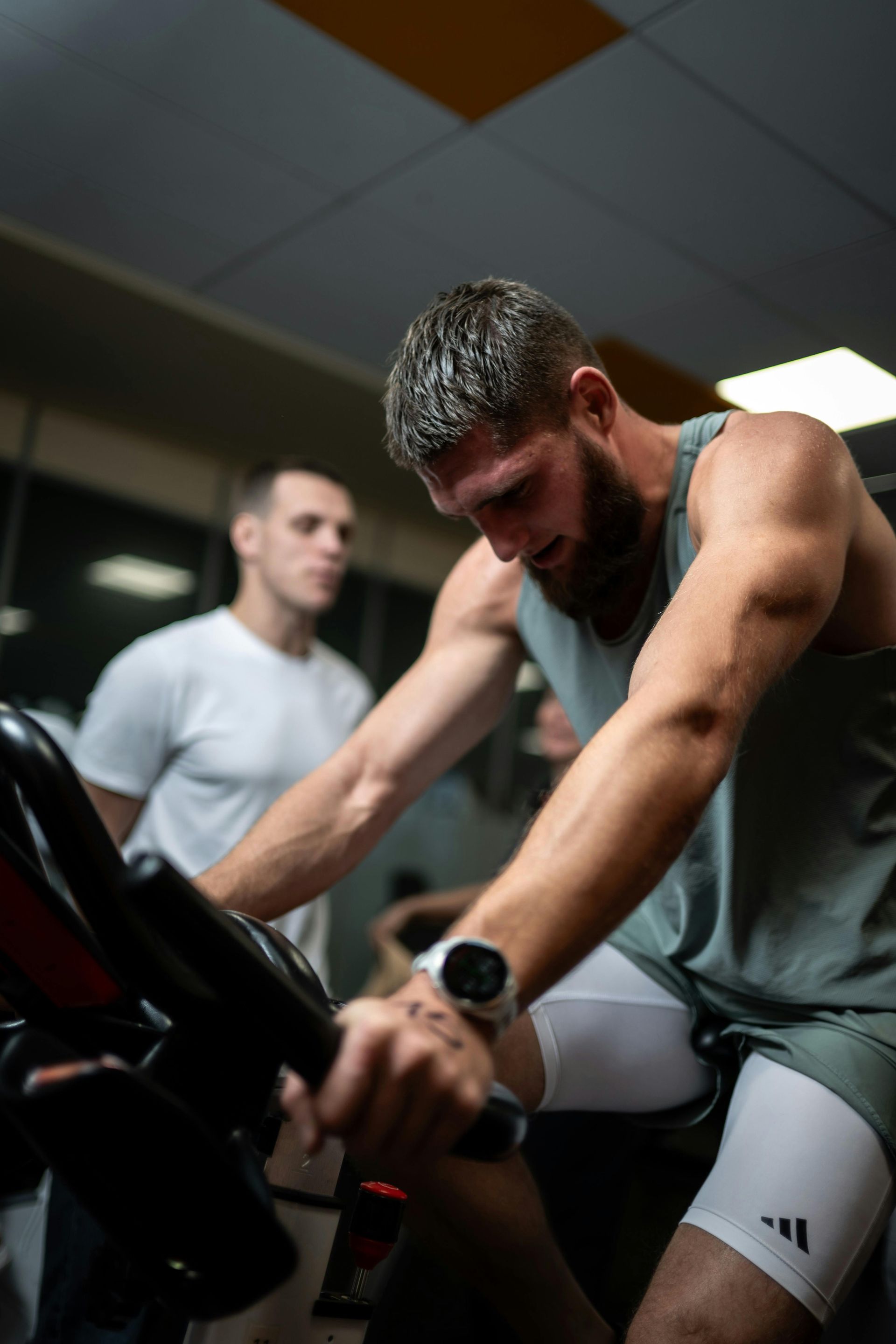 Man pedaling on a stationary bike in a gym, wearing a gray tank top and white shorts.