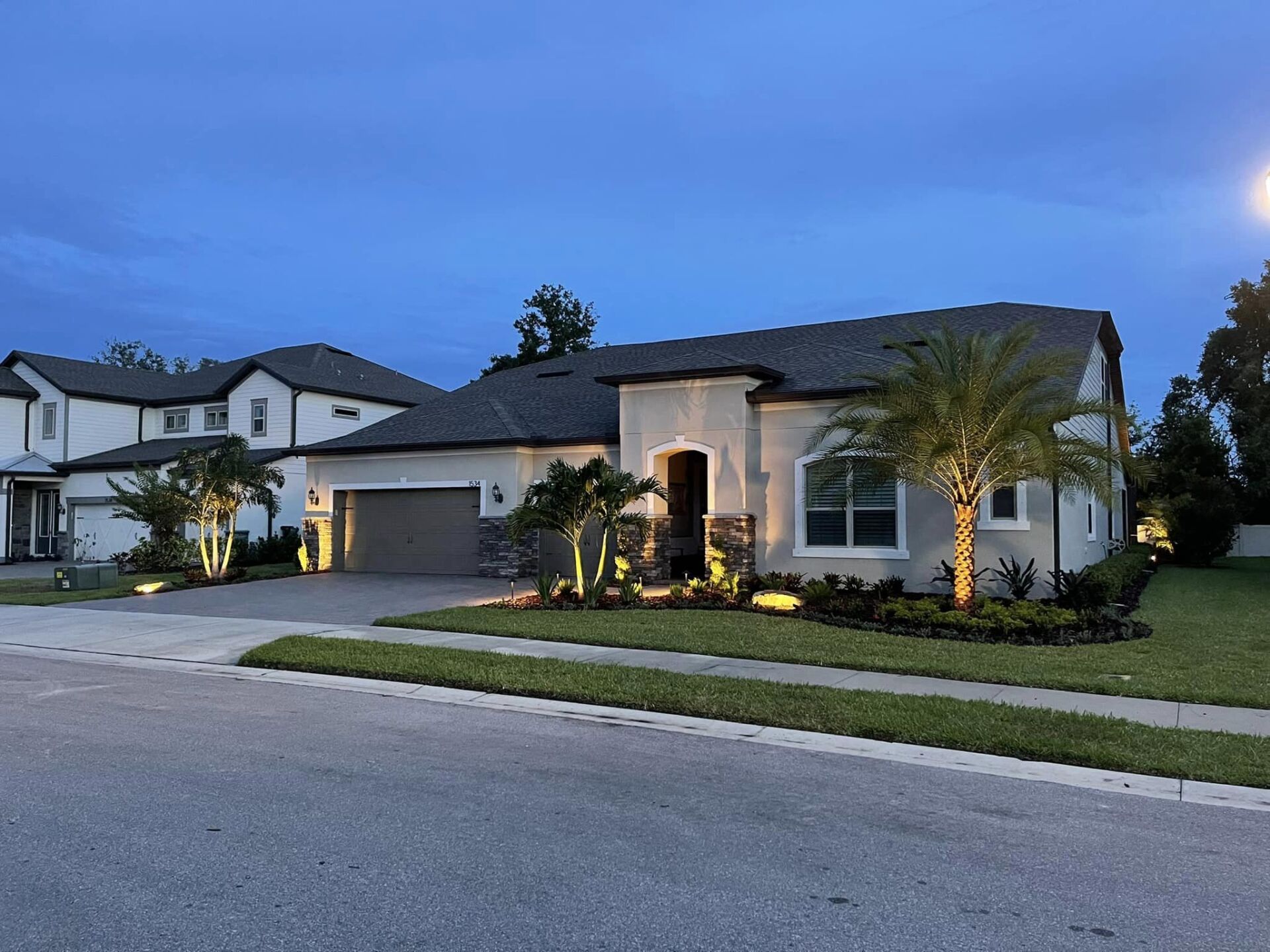 A house with architectural lighting at dusk; beige exterior, garage, palm trees, and manicured lawn.