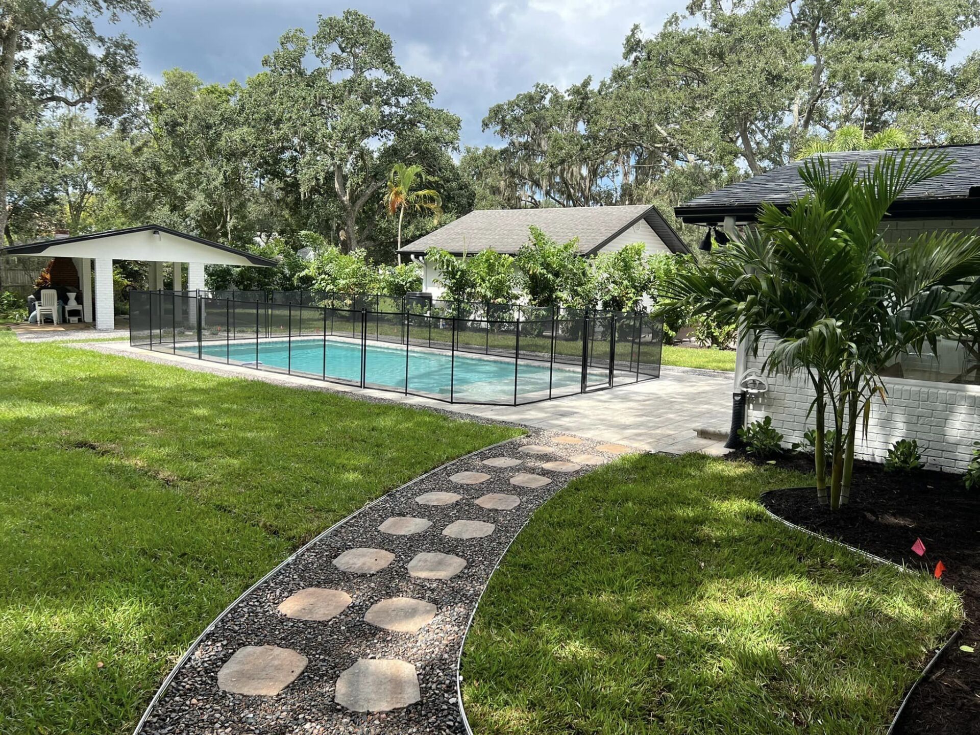 Stone path leads to a backyard pool area with a white cabana, surrounded by lush greenery.