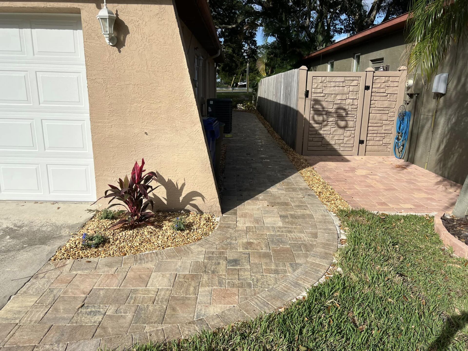 Brick pathway alongside a building, leading to a brown gate. Landscaping and trash can visible.