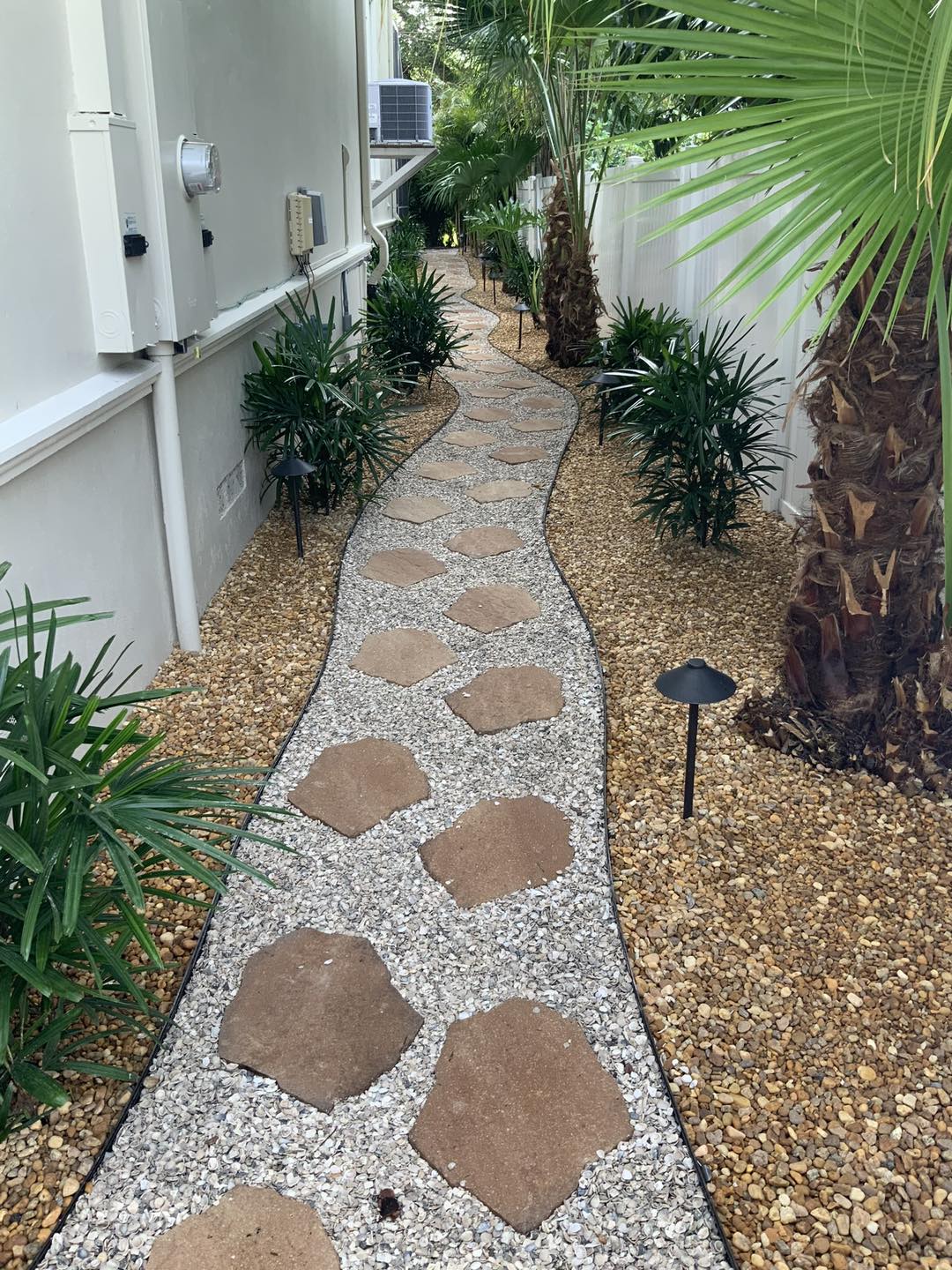 Stone path winding between a white building and lush greenery. Gravel, brown stones, and palm trees.