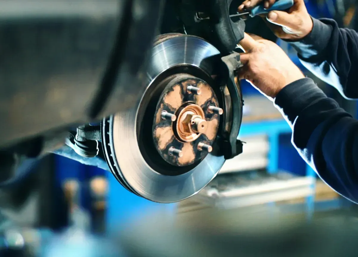Mechanic working on a car brake; hands visible. Shiny rotor, calipers, and suspension. Blue background.