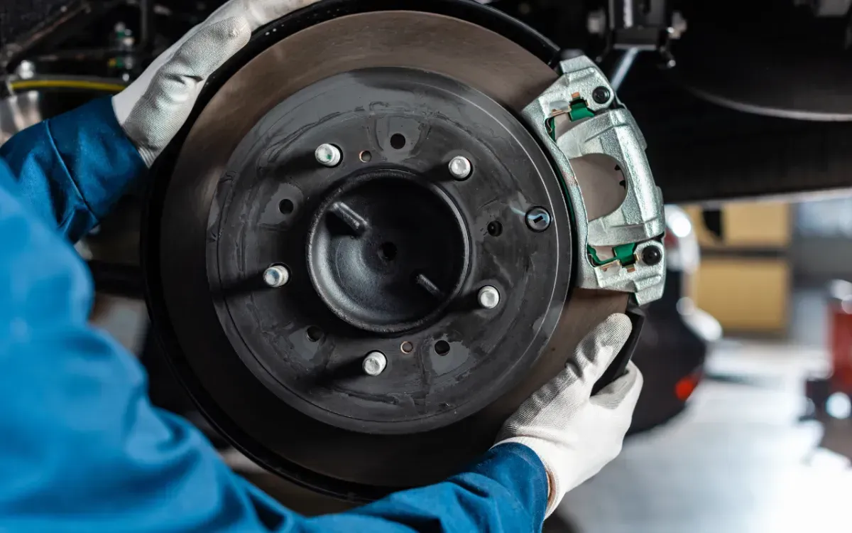 Mechanic holding a car brake rotor and caliper while working on a vehicle in a garage.