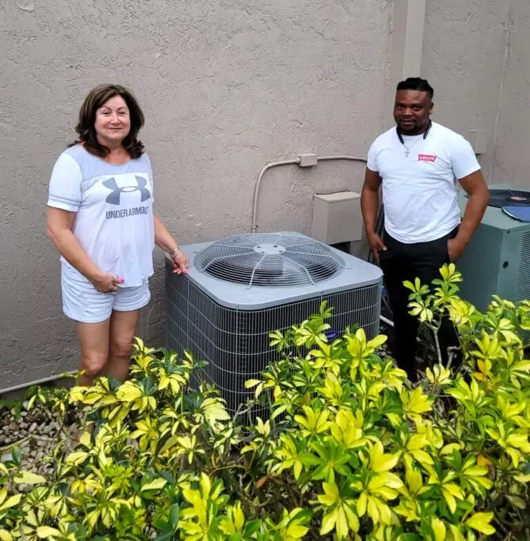 Woman and man standing by an air conditioning unit outside a building, surrounded by plants.