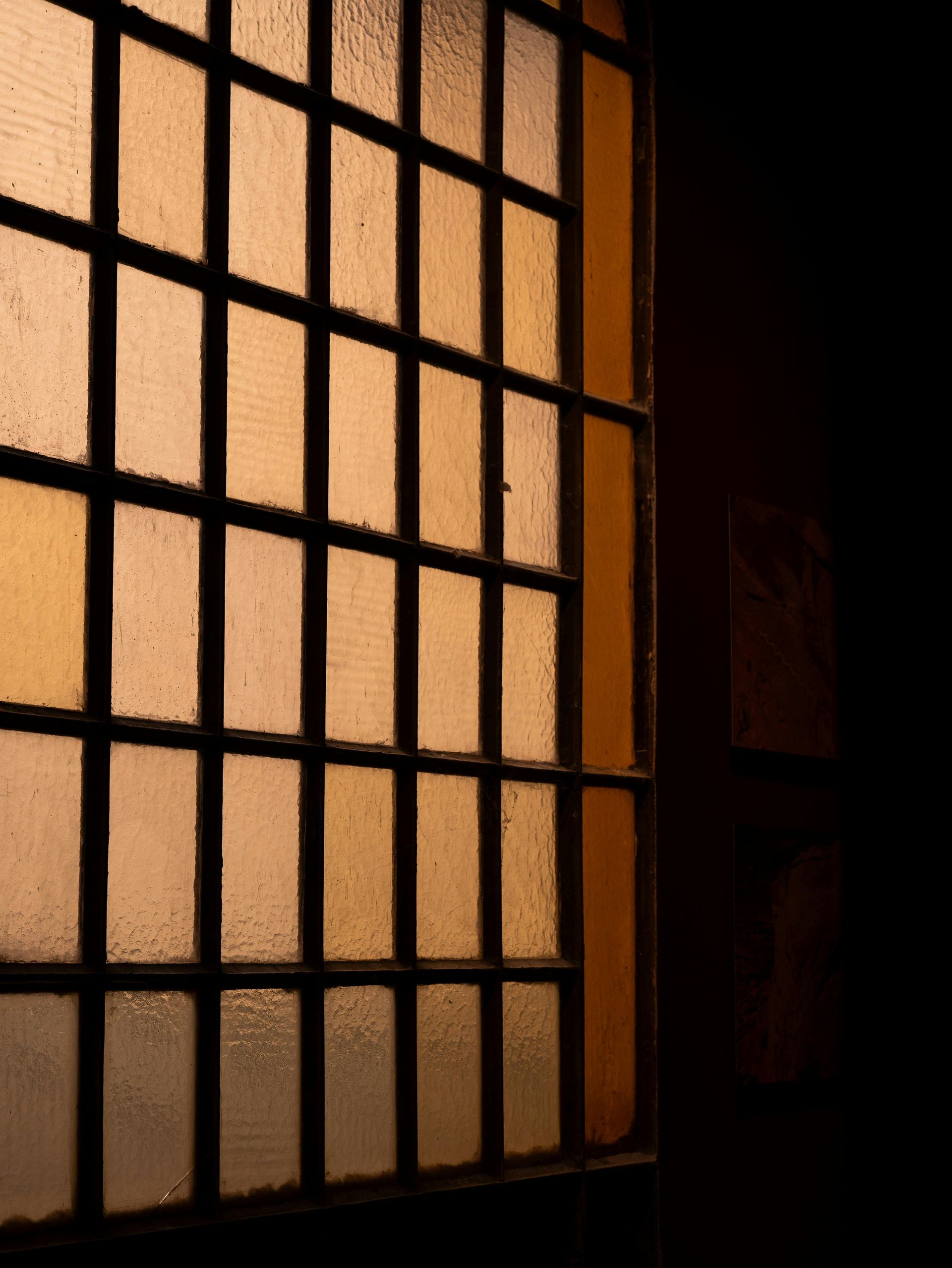 A barred window with yellow and orange light shining through, casting shadows on a dark wall.