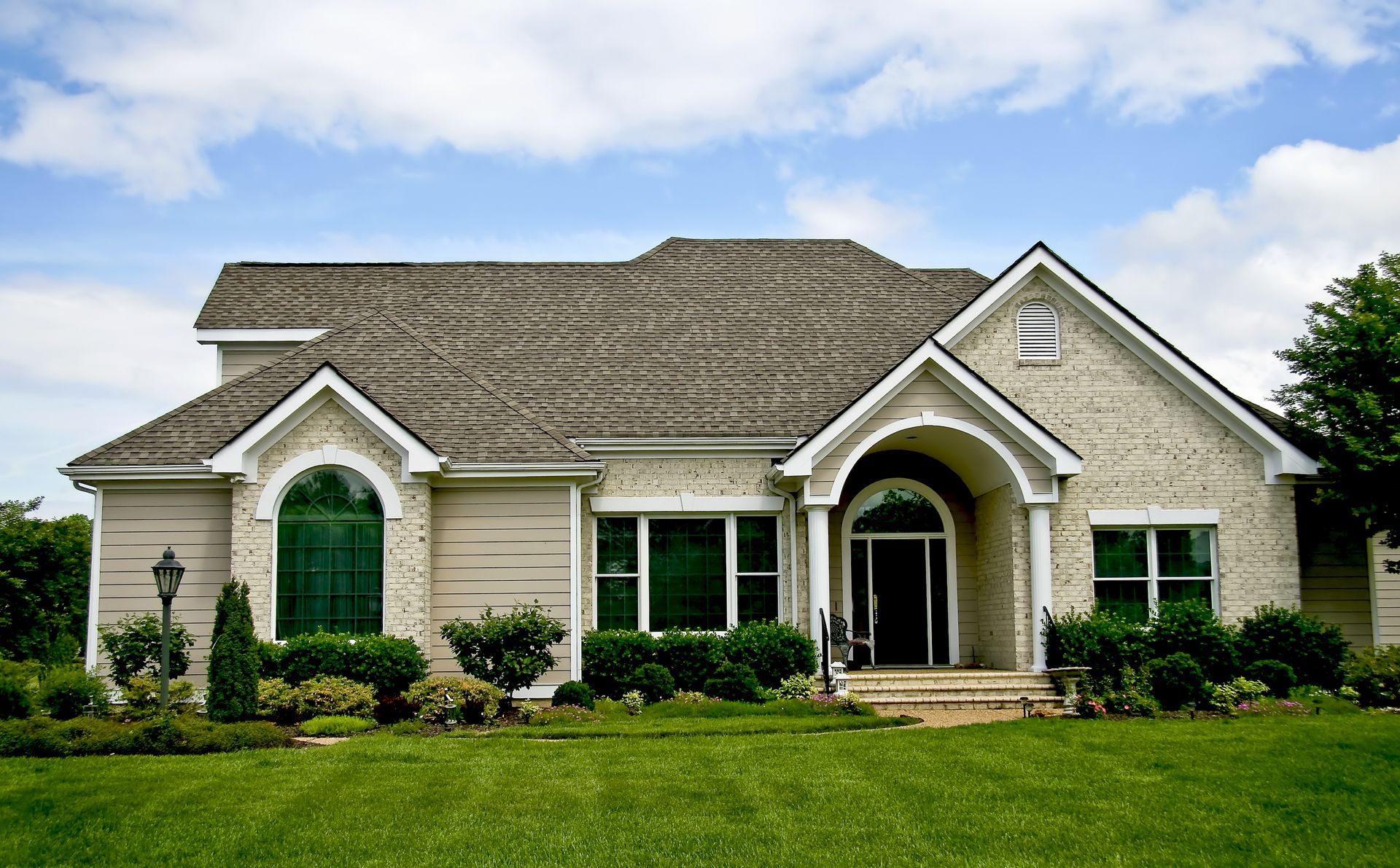 House with tan brick exterior, green windows, and lush green lawn under a blue sky.