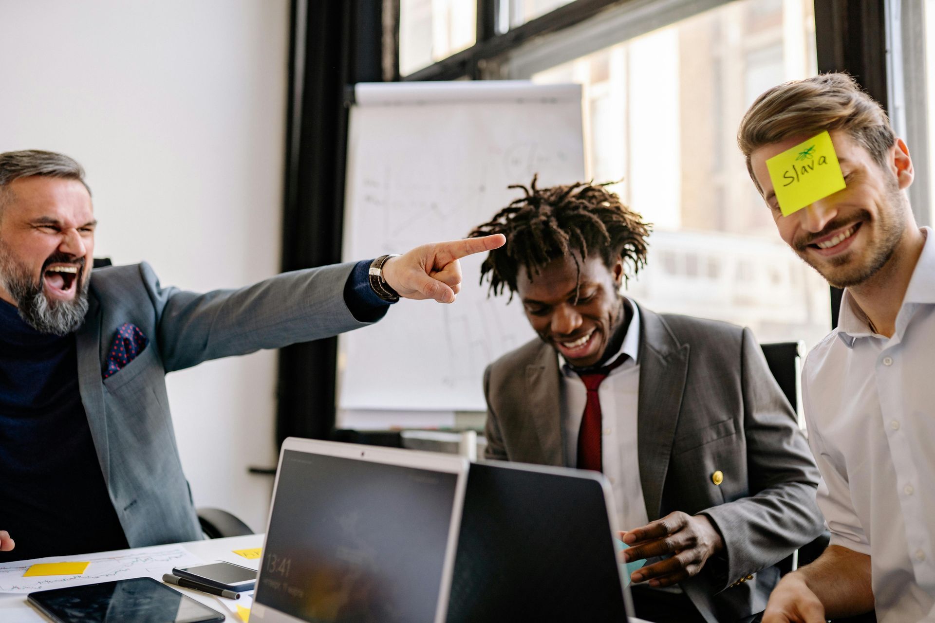 A man is pointing at another man with a sticky note on his forehead.