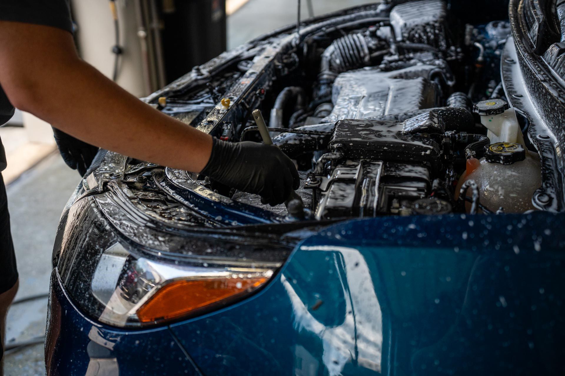 Person cleaning a blue car engine with soapy foam, wearing black gloves.