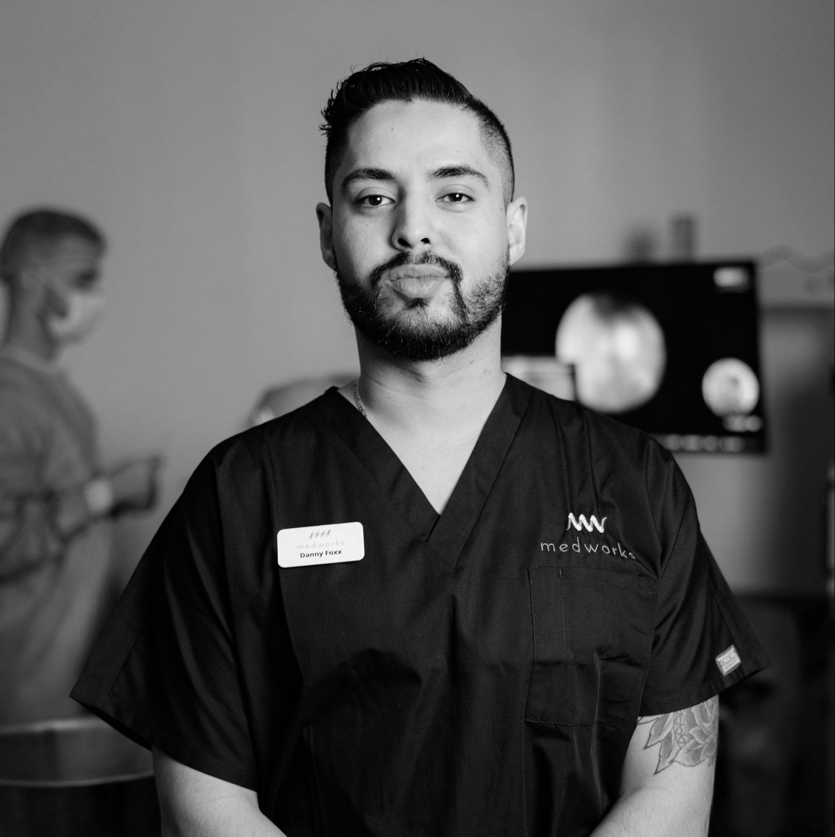 A man with a beard is wearing a scrub top and a name tag.