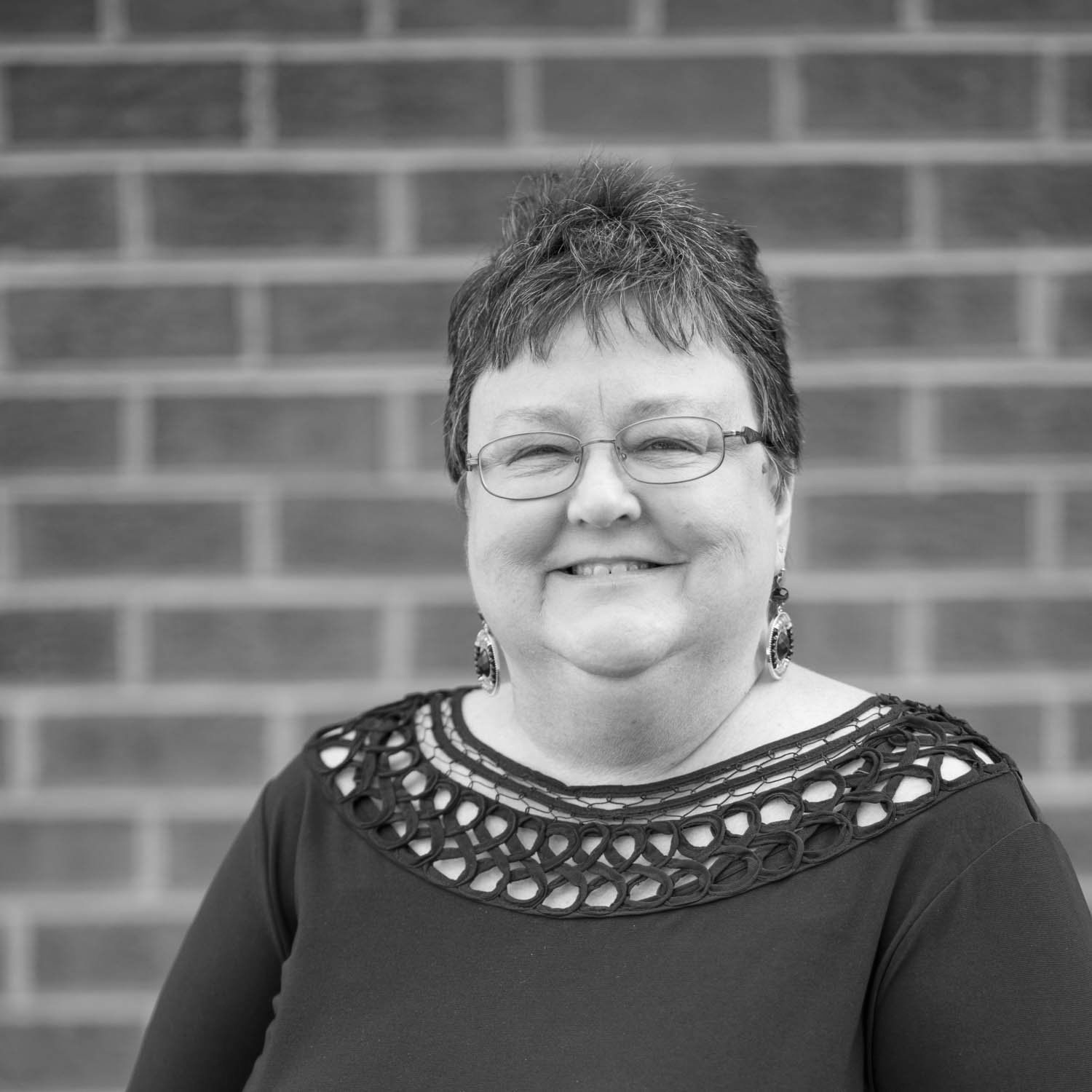 A woman wearing glasses and earrings is smiling in front of a brick wall.