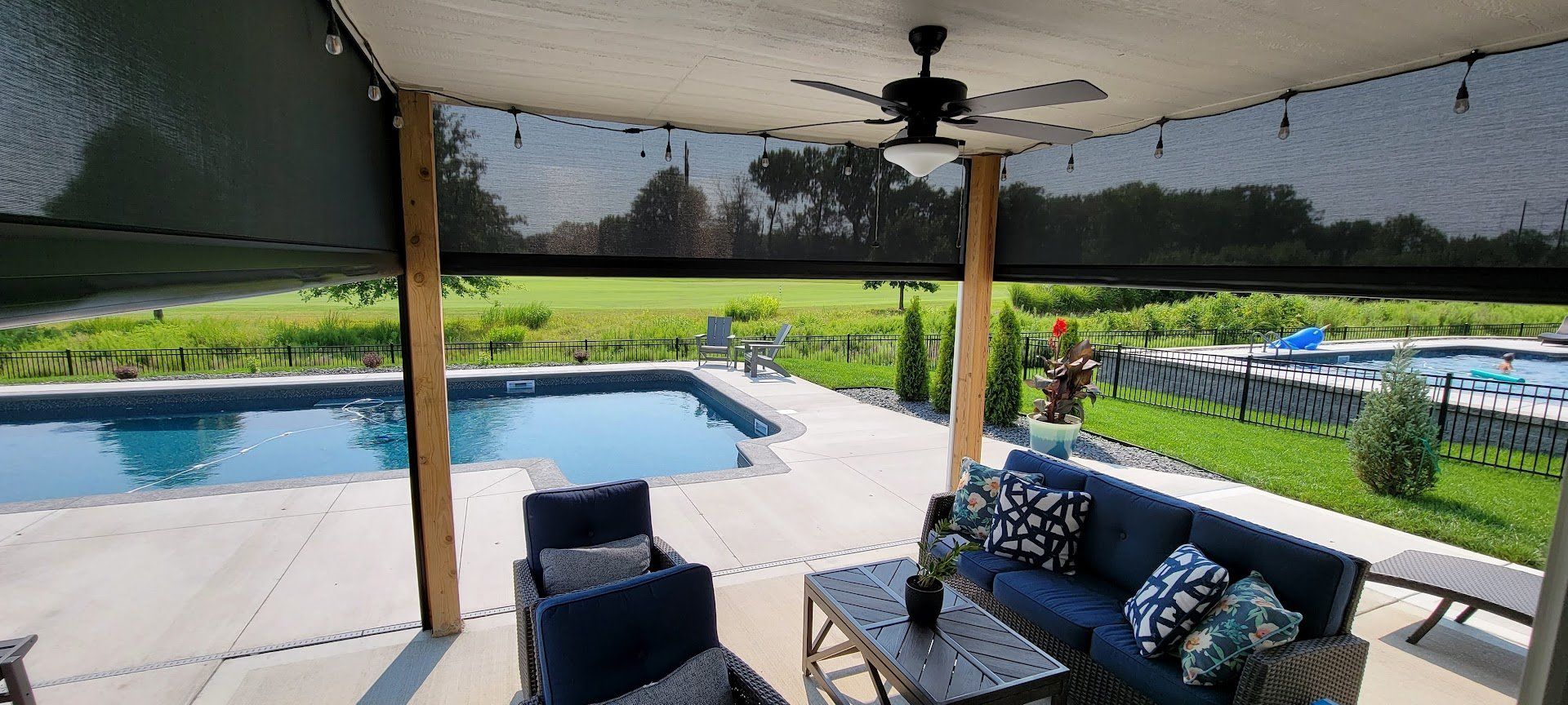 A patio with a couch , chairs , table and a ceiling fan overlooking a pool.