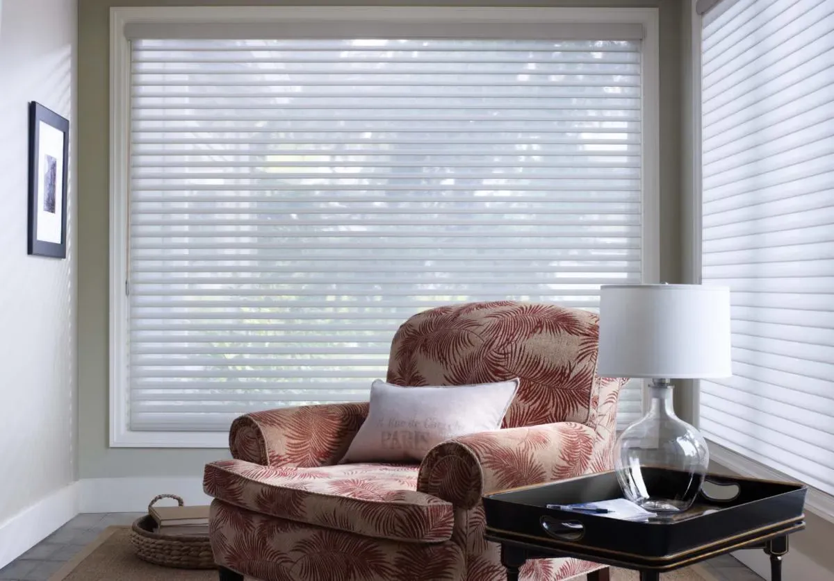 A living room with a chair , table , lamp and window with blinds.