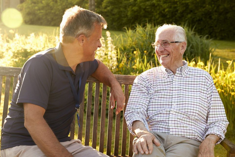Two men sitting on a park bench, smiling and talking in warm sunlight.