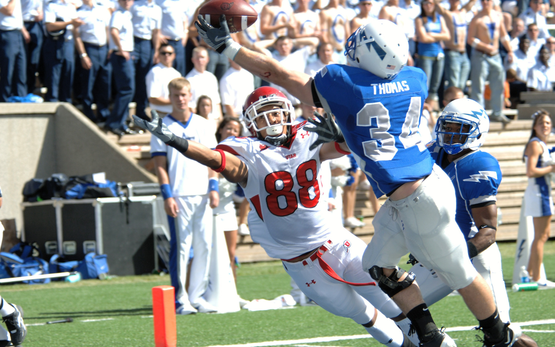 Football player in red leaping to catch a pass, pursued by a blue-clad defender. Stadium crowd in the background.