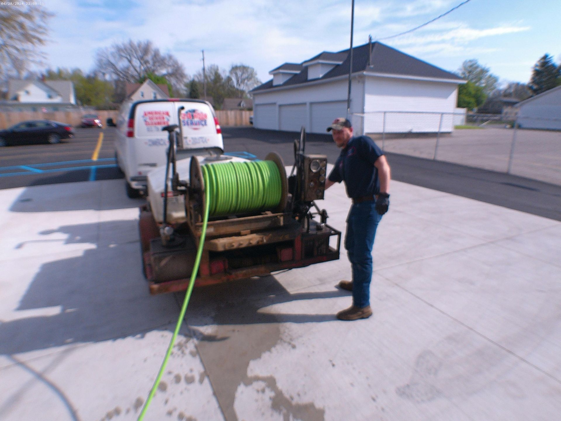 A man operating a green hose reel on a trailer attached to a van, in a parking lot.