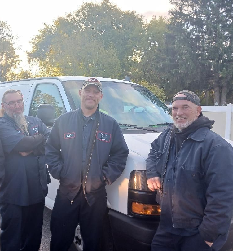 Three men in work clothes stand in front of a white van, smiling.