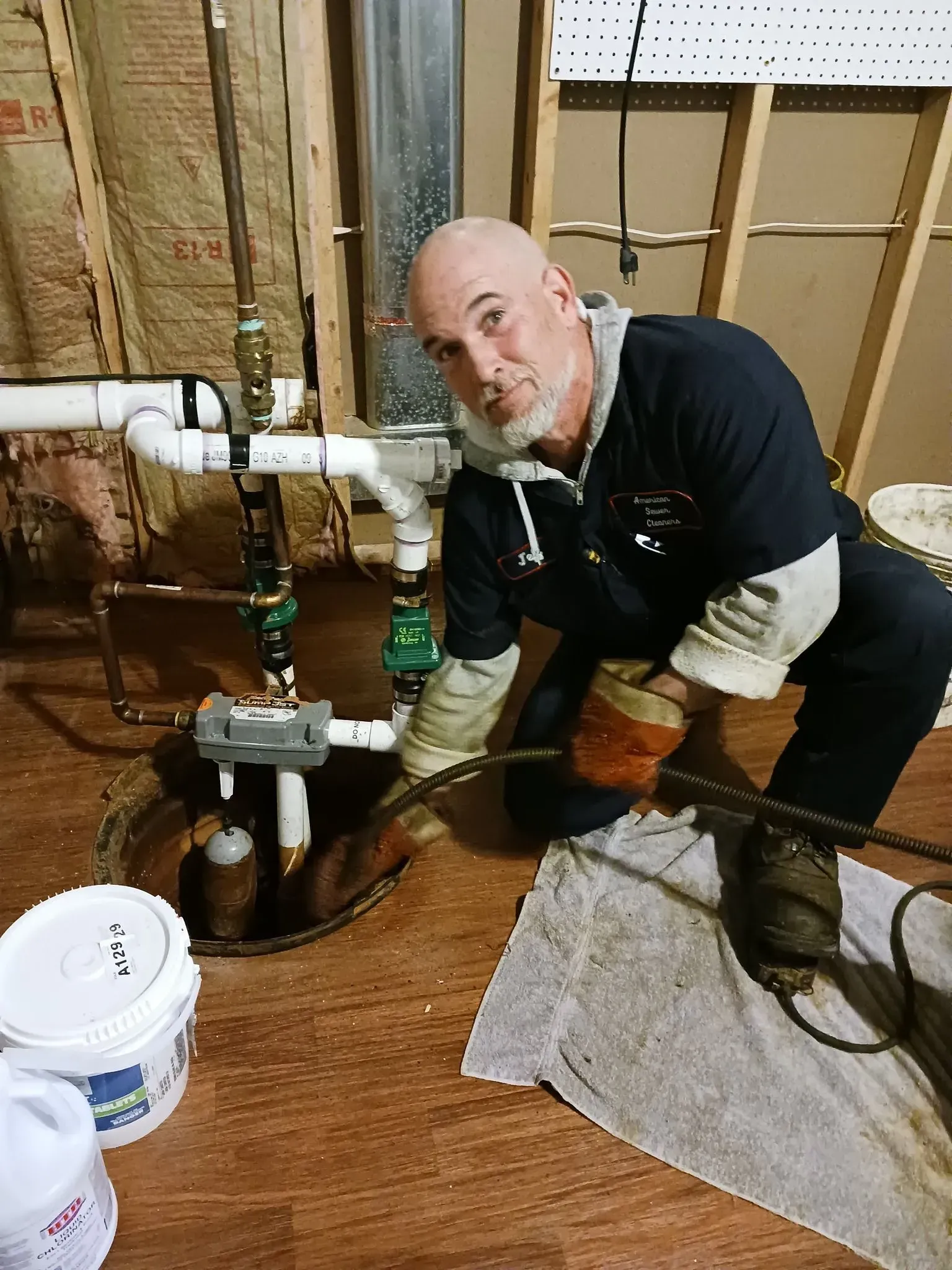 A man working on plumbing, kneeling next to pipes and a pump.