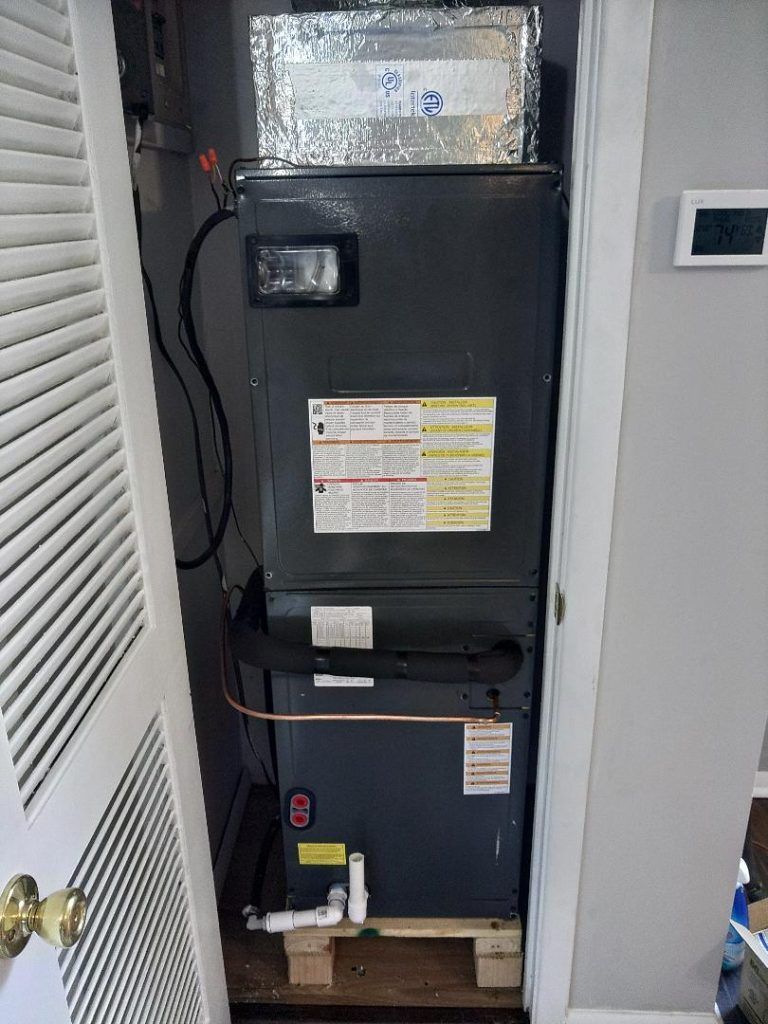 HVAC unit in a closet. Gray metal unit on a wooden base with ductwork and piping. Door open.