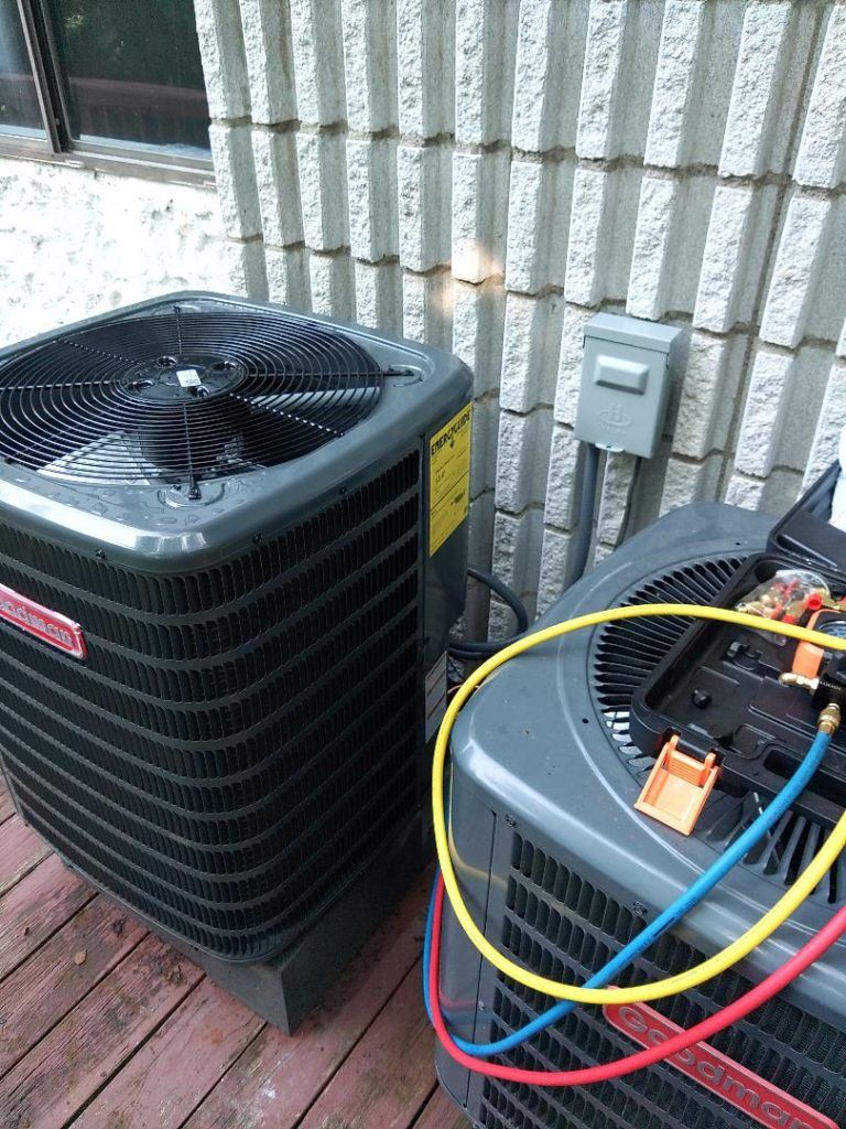 Two Goodman air conditioning units outside, one being serviced with gauges and hoses.
