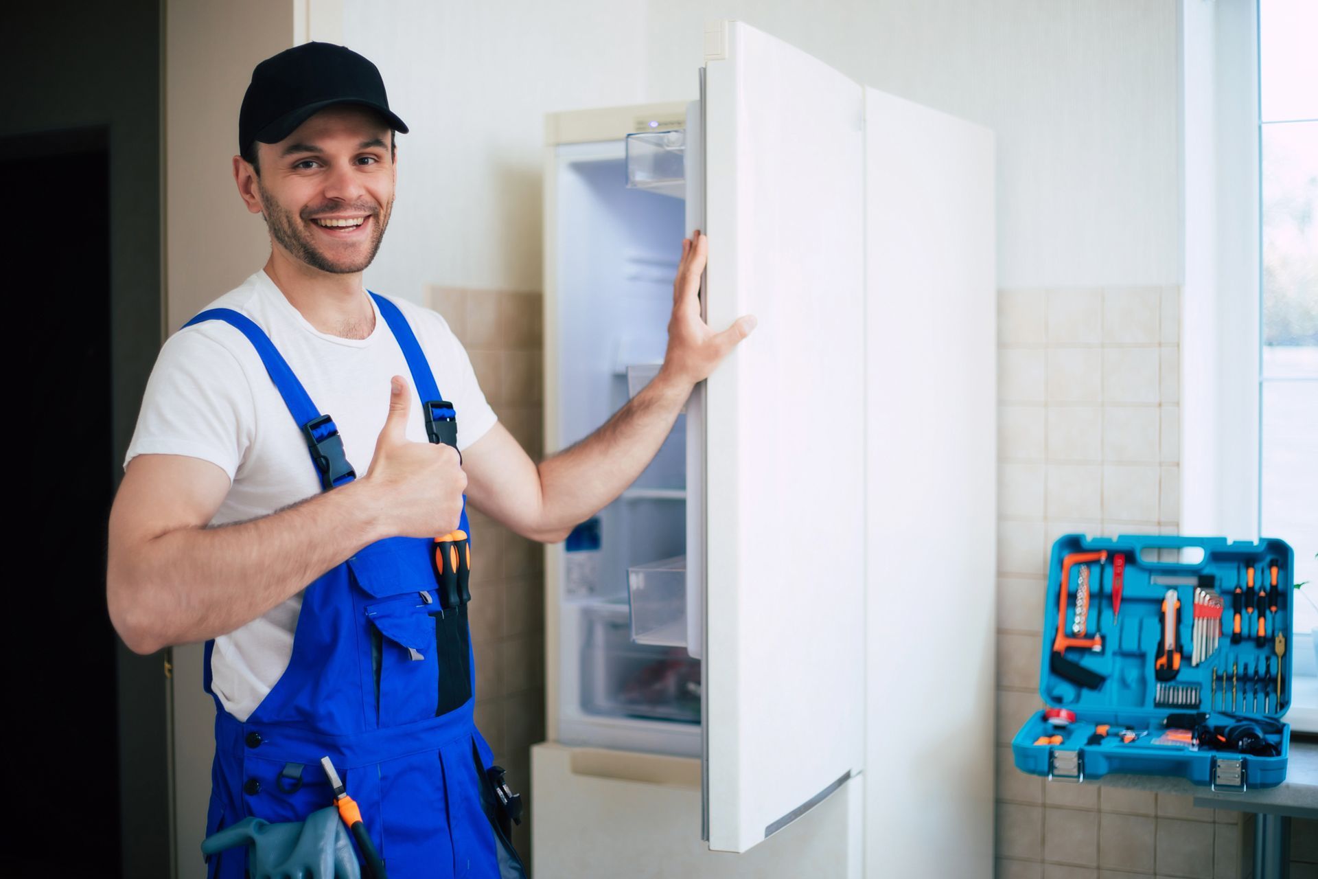 Repairman in blue overalls gives thumbs up beside open refrigerator in kitchen.